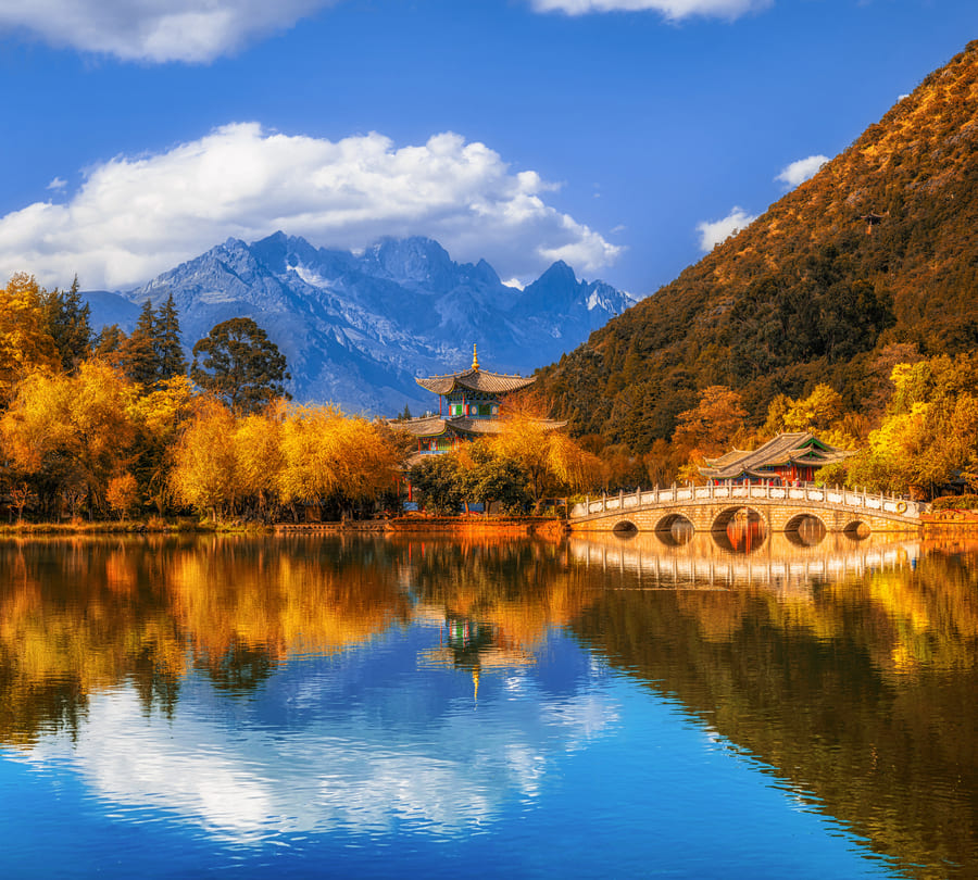 Postal de Yunnan de medio día con lago haciendo efecto espejo, con montañas nevadas de fondo.